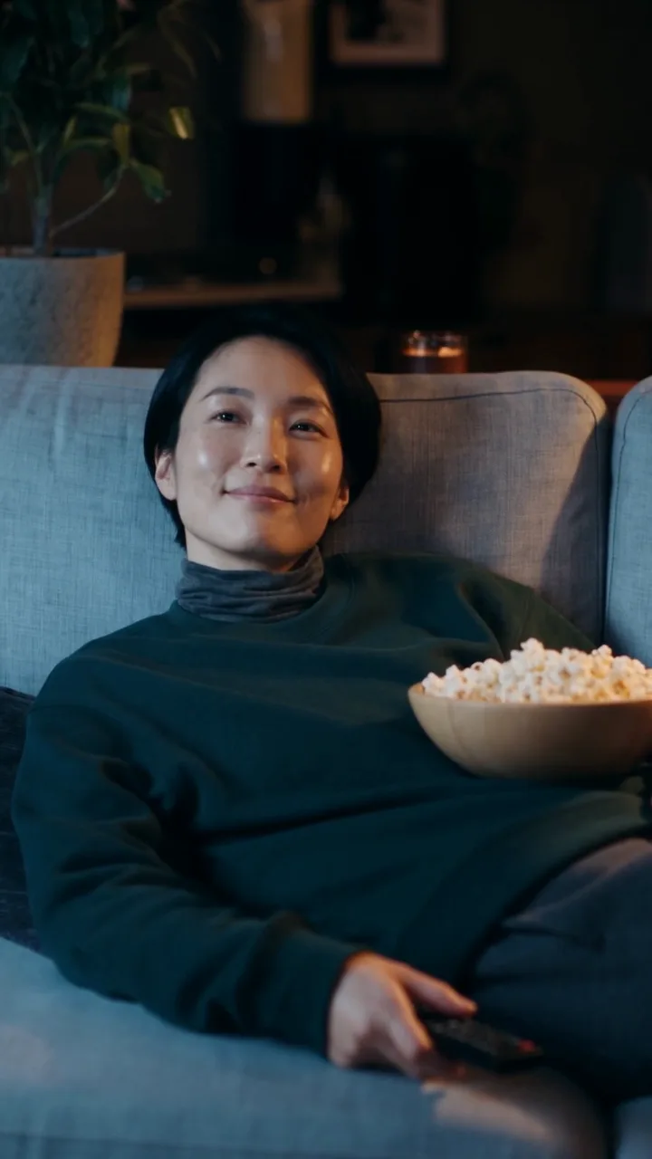 La même femme allongée sur un canapé gris dans son salon, un bol de popcorn posé sur elle, la télécommande à la main.